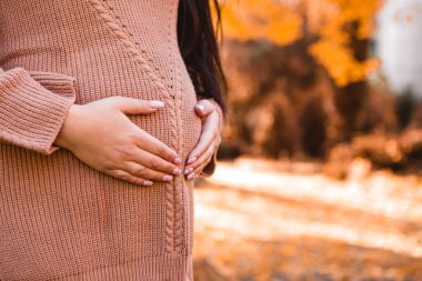 Cropped image of pregnant woman standing in autumn city park, showing heart sign love positive gesture symbol on her round belly with baby child inside. Fall time, red orange and yellow leaves