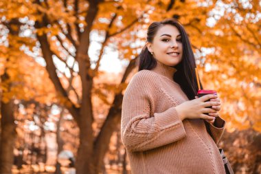 Pregnant woman standing in autumn city park forest, stroking her round belly with baby child inside, drinking hot beverage coffee tea. Fall time. Mother's love, pregnancy concept