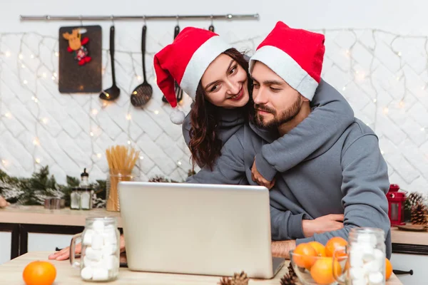 Couple sitting kitchen use laptop buy gifts Internet garlands home cosy interior atmosphere New Year red hats Christmas tree decorations holiday party celebrating greetings concept winter evening
