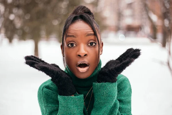 Shocked surprised amazed african american woman standing street outside with white snow background, looking at the camera. New Year atmosphere, winter vacation, cold weather