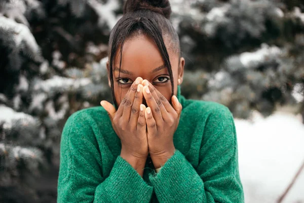 Freezing cold african american woman standing street outside near Christmas Tree, warm her hands with steam form mouth, wear warm mittens. New Year atmosphere, winter vacation, cold weather