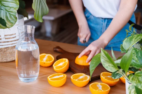 Woman prepares drinking water with fresh oranges at home full of plants. Step by step instruction.