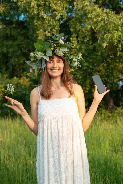 Woman with a flower wreath on her head at the midsummer festival. Holding a mobile phone for shooting. Vertical photo.