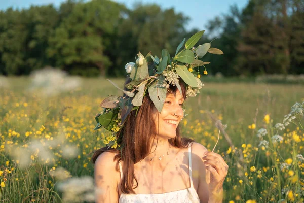 Woman with a flower wreath on her head sits in a field and blows on a dandelion. Summer solstice day. Midsummer festival.