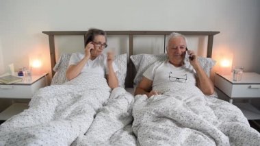 Yaşlı evli çift, erkek ve kadın, akıllı telefon tebessümünü kullanarak evde yatakta dinleniyorlar.