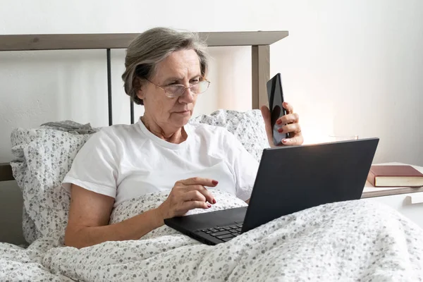 Yaşlanan yaşlı bir kadın yatakta, düz dairede çalışıyor. Ev bilgisayarında Emekliler için akıllı telefon işleri yapıyor..
