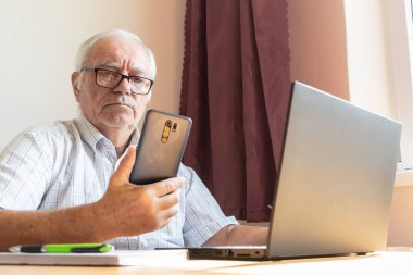 Ana konu odak dışı, kıdemli yaşlı erkek masada oturuyor, ev bilgisayarı çevrimiçi dizüstü bilgisayar iş mesafesi, akıllı telefon.