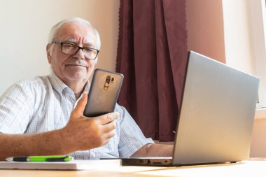 Ana konu odak dışı, kıdemli yaşlı erkek masada oturuyor, ev bilgisayarı çevrimiçi dizüstü bilgisayar iş mesafesi, akıllı telefon.