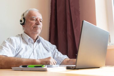 Ana konu odak dışı, kıdemli yaşlı erkek masa başında oturuyor, ev bilgisayarı, dizüstü bilgisayar iş mesafesi serbest müzik kulaklıkları mutlu çağrı merkezleri.