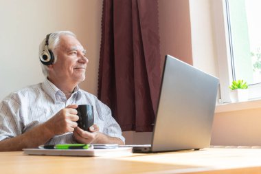 Ana konu odak dışı, kıdemli yaşlı erkek masa başında oturuyor, ev bilgisayarı, dizüstü bilgisayar iş mesafesi serbest müzik kulaklıkları mutlu çağrı merkezleri.
