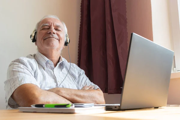 Ana konu odak dışı, kıdemli yaşlı erkek masa başında oturuyor, ev bilgisayarı, dizüstü bilgisayar iş mesafesi serbest müzik kulaklıkları mutlu çağrı merkezleri.