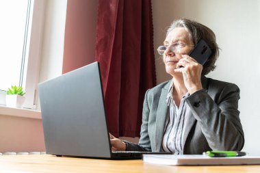 Ana konu odak dışı, yaşlı yaşlı kadın masa başında oturuyor, ev bilgisayarı çevrimiçi dizüstü bilgisayar iş mesafesi serbest, akıllı telefon.