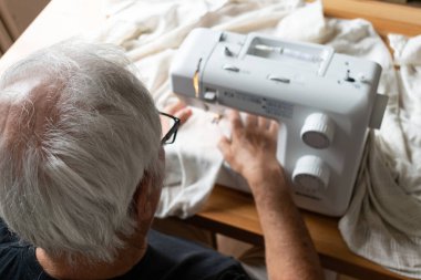 the main subject is out of focus, old male man senior sit next at table at home work, sewing machine enjoy happy relax and make money