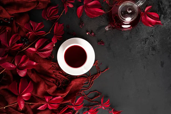 Herbstliche Komposition. Karkadetee in einer Teekanne, eine weiße Tasse mit Untertasse, ein Schal und herbstliche rote Traubenblätter. Flach lag er. Ansicht von oben. Stockbild