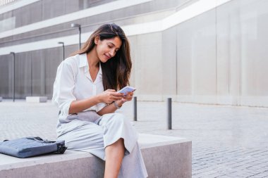 Genç Hintli kadın iş kadını bankta oturuyor ve akıllı telefonunu kullanıyor.