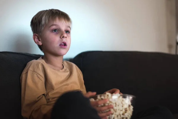 Enfant curieux manger du pop-corn tout en regardant la télévision avec intérêt sur le canapé Image En Vente