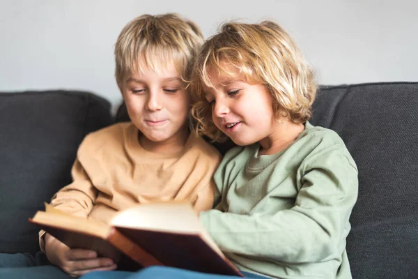 Niños positivos leyendo interesante libro en el sofá y sonriendo — Foto de Stock