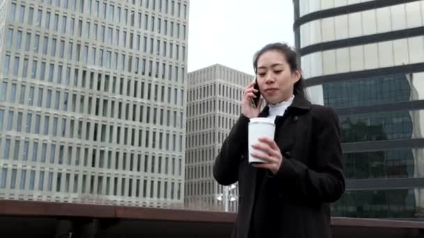 Cheerful Asian businesswoman with coffee to go talking on smartphone — Stock Video