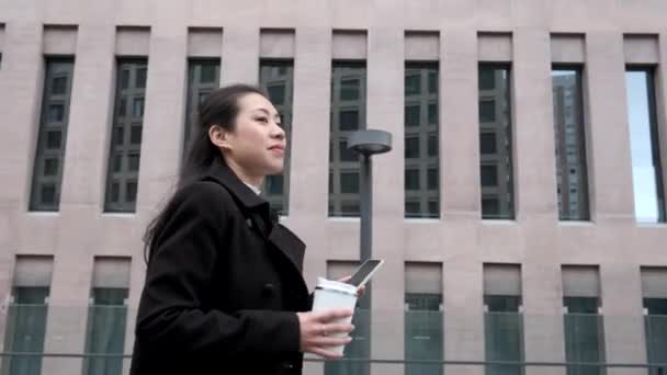 Азиатская бизнесвумен с сотовым телефоном и кофе пойдет гулять по улице — стоковое видео