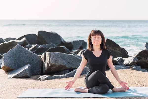 Full body of concentrated flexible female sitting in Gomukhasana with closed eyes and practicing yoga on sandy coast in summer