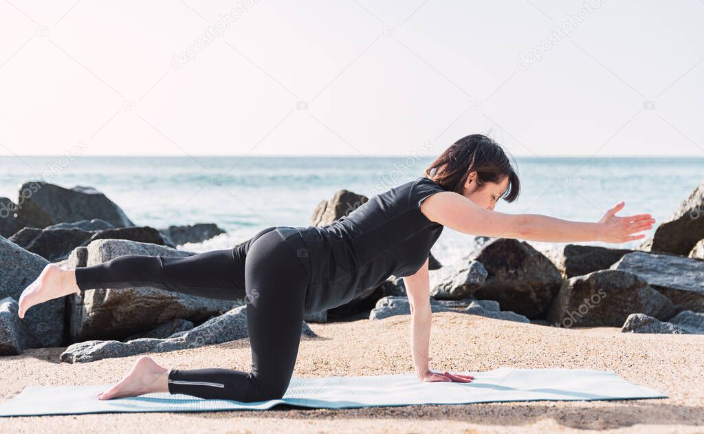 Full body side view of barefoot female in activewear balancing on mat in Chakravakasana while practicing yoga on sandy beach against sea in summer