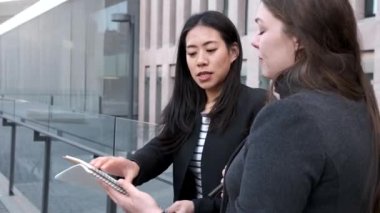 Multiethnic businesswomen reading notes in notepad and talking about new ideas while standing in street and looking at each other