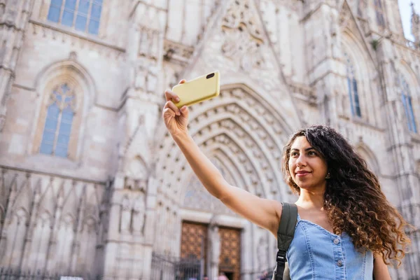 Low angle of young Hispanic woman with long curly hair taking selfie outside medieval cathedral on sunny summer day
