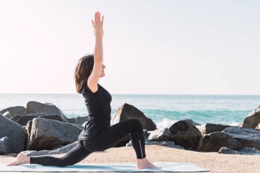 side view of serene female in sportswear. She is stretching legs in Anjaneyasana while practicing yoga on rocky beach against sea in sunny summer day