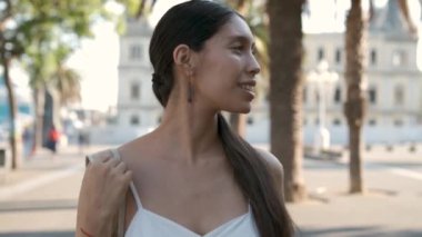 Tracking shot of cheerful Latin American woman with bag smiling and looking around while strolling near palms on sunlit street of Barcelona, Spain