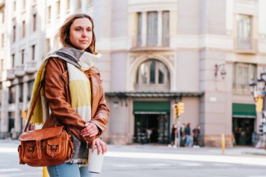 Sokakta toplu taşıma bekleyen trend sarışın bir kadının yatay portresi. Elinde yeniden kullanılabilir bir kahve fincanı var.