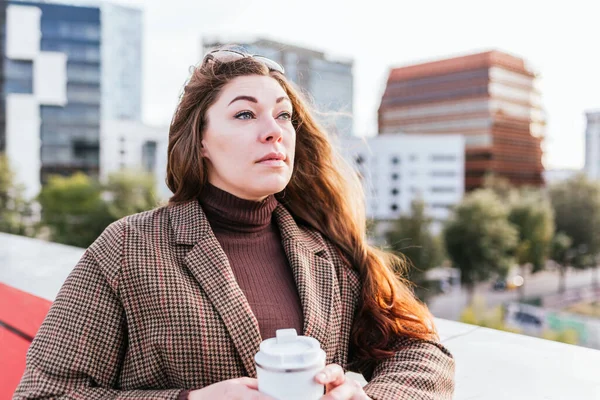 Thoughtful trendy woman with zero waste cup of takeaway coffee looking away while resting on blurred background of city street in daytime