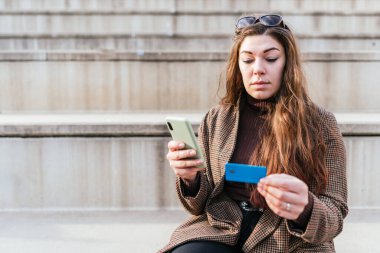 Kareli ceketli bir kadın kredi kartı bilgilerini cep telefonuna girerken merdivenlerde oturuyor ve şehirde internetten sipariş veriyor.
