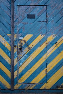 Mavi ve sarı ahşap ev kapısı. Yıpranmış, yaşlanmış, pejmürde, klasik bir giriş. Boyalı yüzey dokusu arkaplanı soyulmuş. Çapraz çizgiler. Hasarlı metal kilit. Dikey