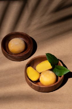 Hafif arka planda Mochi dondurması. Ahşap bir kasede mango meyveli tatlı. Geleneksel Japon tatlıları. Tropik, güneşli ve palmiye yapraklı bir poster. Yaz havası. Dikey, yakın, seçici odaklanma