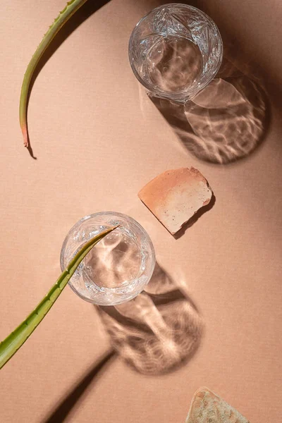 Glass with water in sunshine, light beige background. Summer gin cocktail with aloe vera and lime. Aesthetic, natural refreshing drink concept. Harsh sun shadow. Hot, mexican vacation vibe