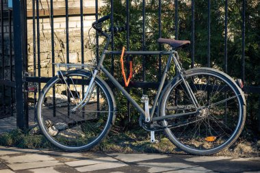 Black retro bicycle parked against metal fence with house wall and garden in the background. Summer, ecological, popular transport. Vintage bicycle with lock. Safe parking in the city