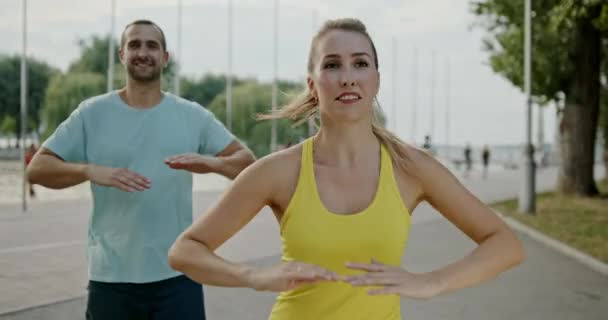 男と女は同時に一緒に肩や腕の上で運動を実行します 夏の日にウォーターフロントで積極的なウォーミングアップ 一緒に訓練は常により効果的です — ストック動画