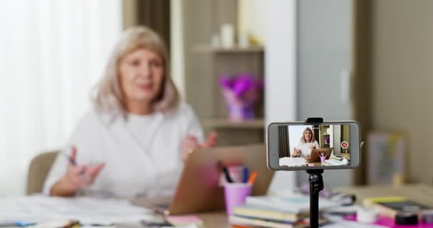 上級教師の録画ビデオレッスン 教育ブログのためのビデオレッスンを記録しながら 文房具やノートパソコンで被写体を説明する高齢女性のラックフォーカス — ストック動画