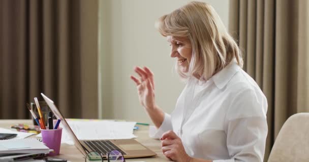 高级自由职业者在家里打视频电话 穿着白衬衫的老年妇女在网上向同事致意 并在家中的虚拟会议上带着文件 — 图库视频影像