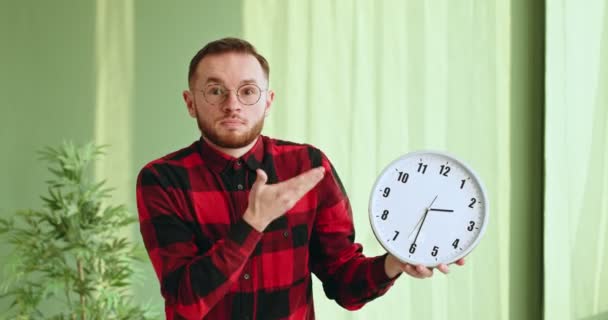 时间管理的概念 年轻的留着胡子的男性穿着随意的格子衬衫 戴着眼镜 指着钟 看着镜头 对时间的延误感到愤怒和不满 — 图库视频影像