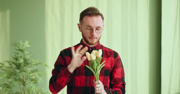 身穿格子衬衫 戴着眼镜 留着胡子的正年轻男性 吹散鲜花中的灰尘 并将花束送给相机 — 图库视频影像