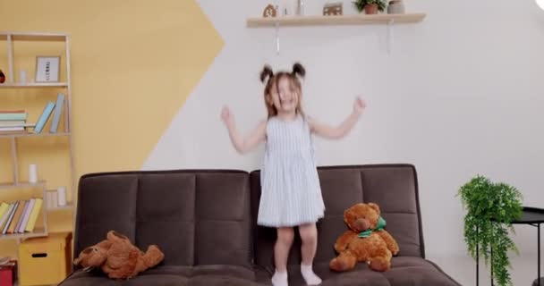 Feliz Niña Vestido Calcetines Levantando Brazos Sonriendo Mientras Salta Sofá — Vídeo de stock