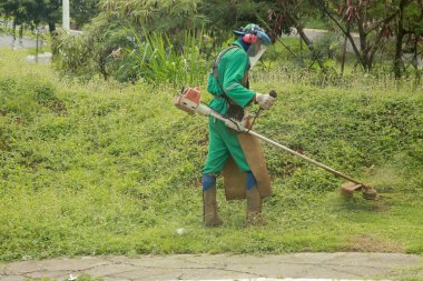 Salvador, Bahia, Brezilya - 24 Kasım 2021: Kamu temizlik işçisi Salvador 'daki kamu yollarında çalı biçmek için bireysel koruma ekipmanı ve çim biçme makinesi kullanıyor.