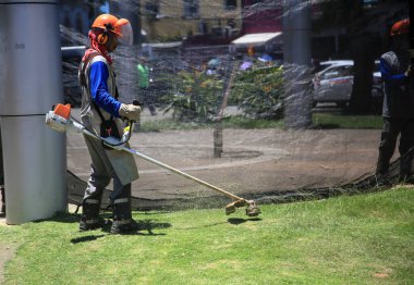 Salvador, Bahia, Brezilya - 12 Ocak 2022: Bir kamu temizlikçisi Salvador şehrindeki bir meydanda çim biçme makinesi kullanarak çim biçiyor..