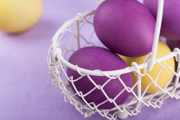 Colored eggs in a white basket on lilac background — Stock Photo, Image