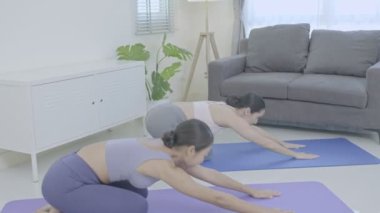 Asyalılar oturma odasındaki bir paspasın üzerinde yoga yaşam tarzı dersi alıyorlar. Spor kıyafetli genç kadınlar evde yoga eğitimi vermeyi öğretiyor..