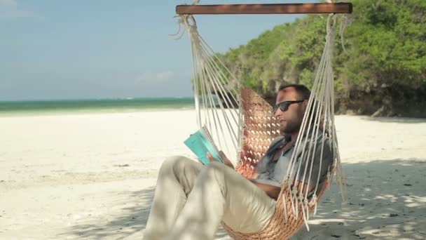 Молодой человек читает книгу, сидя, расслабляясь на гамаке — стоковое видео
