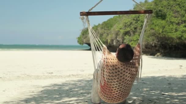 Νεαρός άνδρας, χαλάρωση, ενώ κάθεται σε αιώρα στην πανέμορφη εξωτική παραλία — Αρχείο Βίντεο