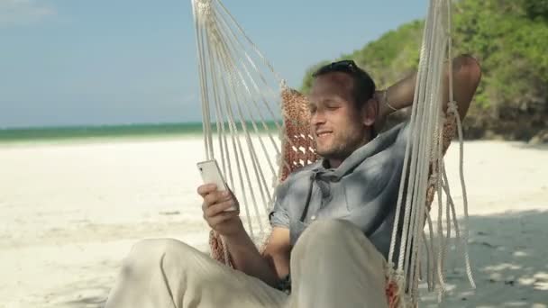 Jonge man op gebruikend smartphone op prachtige exotische strand hangmat — Stockvideo