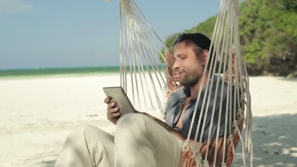 Jeune homme avec tablette sur hamac, plage exotique — Video